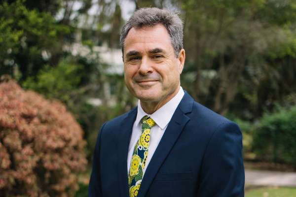 Grant Stanley in a navy suit standing in front of greenery smiling at the camera with his hands folded in front of him